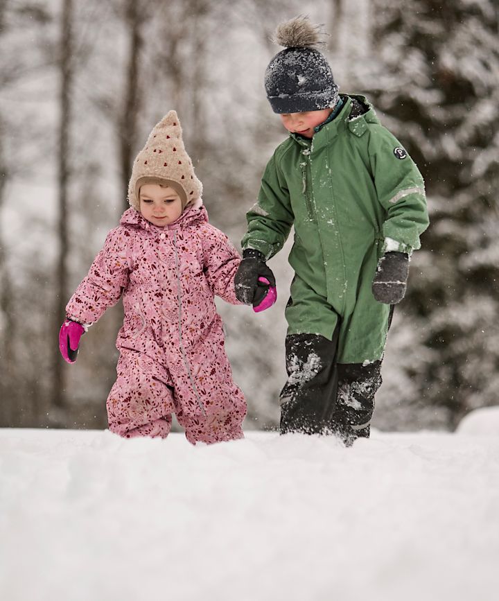To barn i vinterdrakter går hånd i hånd i snøen - en i rosa dress med beige lue og en i grønn dress med mørk lue.