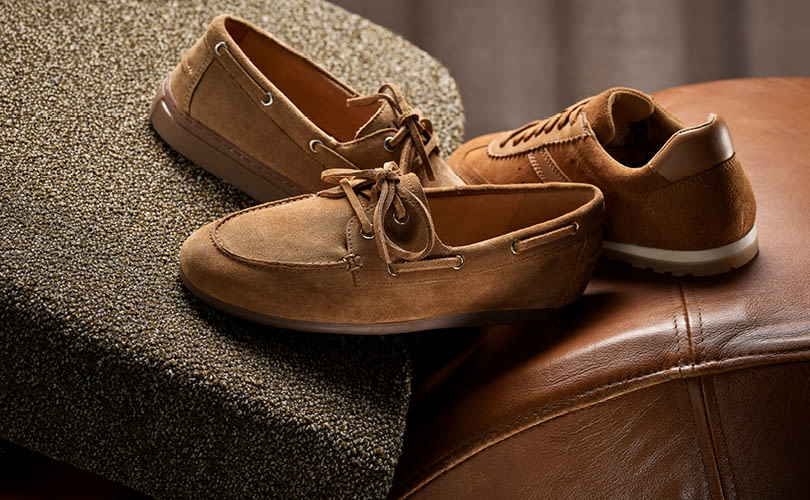 Brune semsket båtsko og casual sneakers i lær plassert på en brun lærsofa og tekstilpute med nøytral bakgrunn.