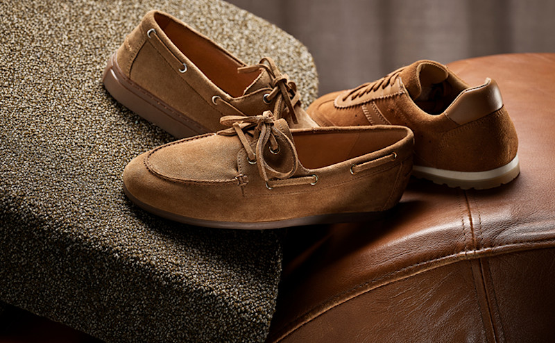 Brune semsket båtsko og casual sneakers i lær plassert på en brun lærsofa og tekstilpute med nøytral bakgrunn.
