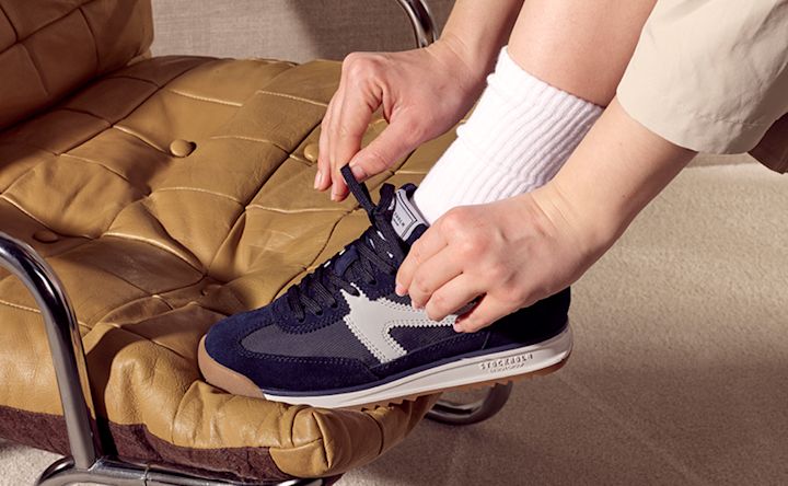 Person knyter snørebåndene på en marineblå retro-sneaker med grå detaljer og hvit sål, mens de sitter i en stol.
