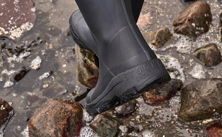 Person med svarte støvler trår på steiner i grunt vann, sålen viser teksten "STOCK DESIGN".