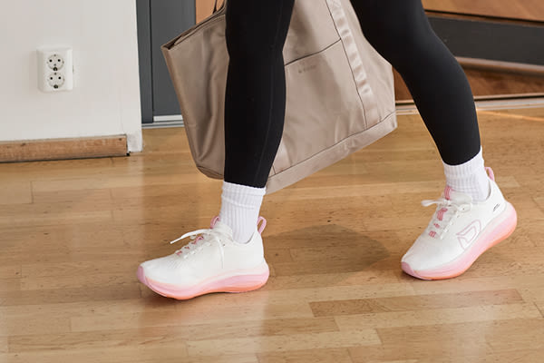 Person med svarte tights og hvite fritidssko med rosa såler fra Okaki bærer en beige veske på tregulv.