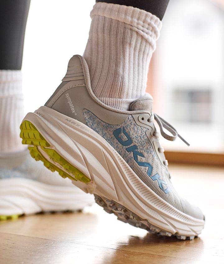 Lyseblå fritidssko fra Okaki med hvit såle, gule detaljer og hvite sokker, vist fra siden på tregulv.