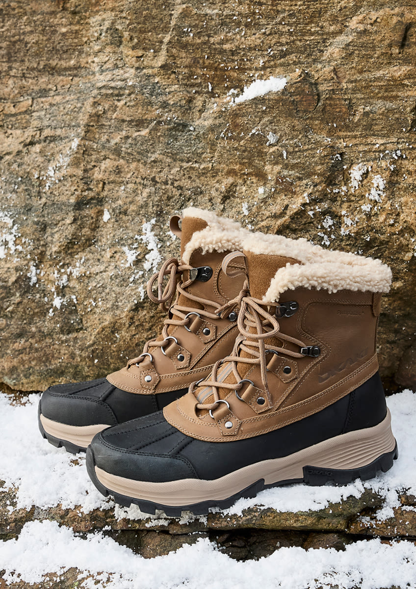 Bruna vinterkängor från Okaki med svart gummisula och fluffig fårskinnskant står på snötäckt stengrund mot en stenvägg.