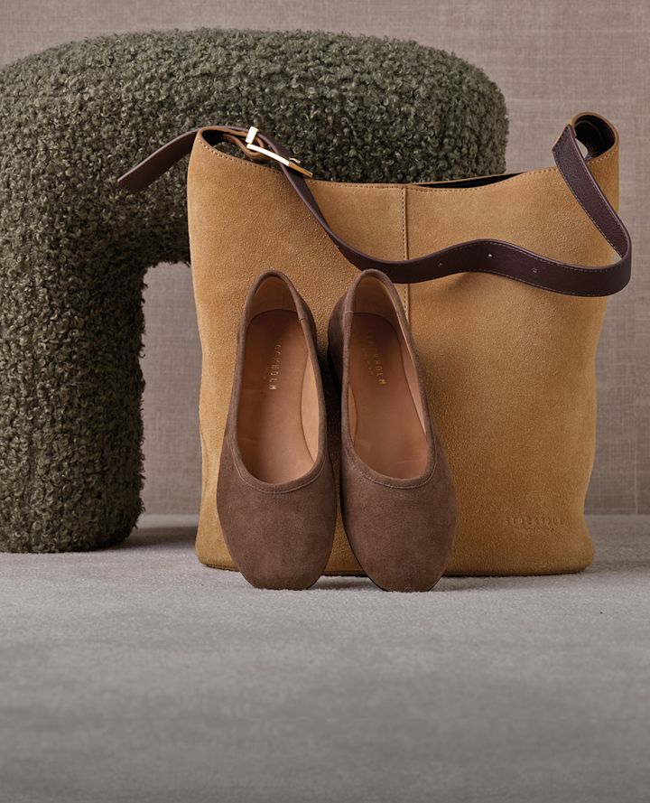 Bruna mockaballerinaskor framför en kamelbeige mockaväska med brunt läderhandtag, placerade på en grå yta bredvid en grön puff.