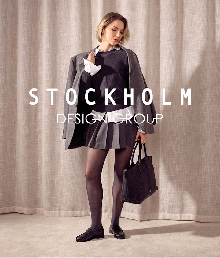 Kvinna i grå dräkt med svarta loafers i klassisk stil, bär svart väska framför beige gardin med Stockholm Design Group-logotyp.