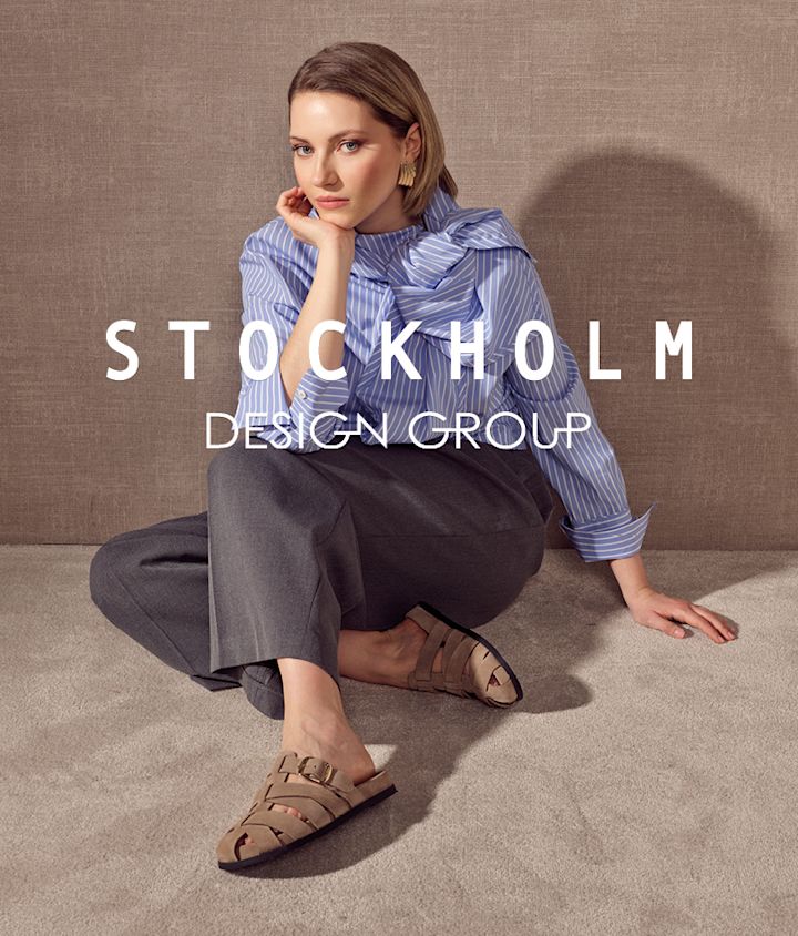 Kvinna sitter på golvet i beige flätade sandaler med spänne, grå byxor och blå randig blus. Stockholm Design Group.