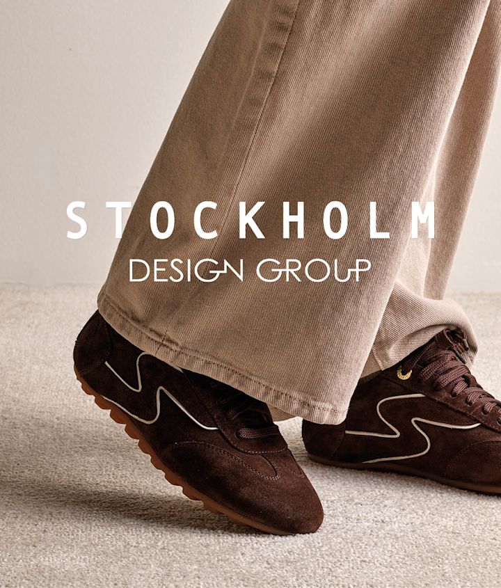 Mörkbruna retro-sneakers i mocka med beige detaljer, parade med beige byxor. Stockholm Design Group-logotyp syns i bild.