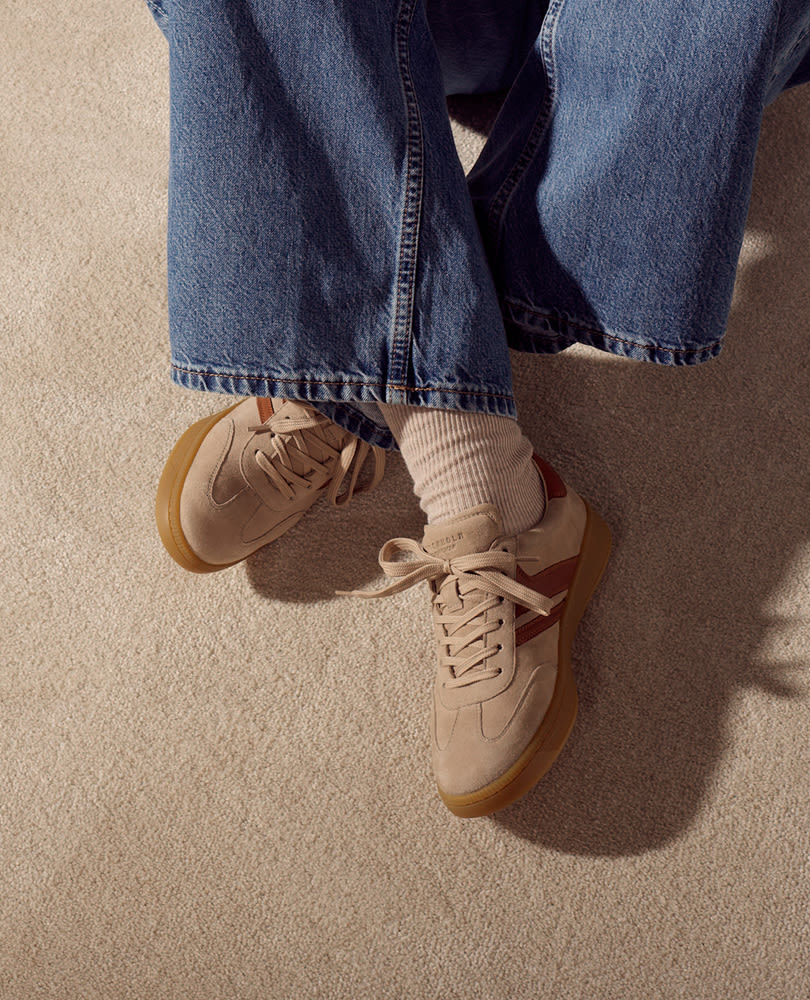 Beige semsket sneakers med gummisåle og røde detaljer, brukt med vide jeans og hvite sokker på beige teppe.