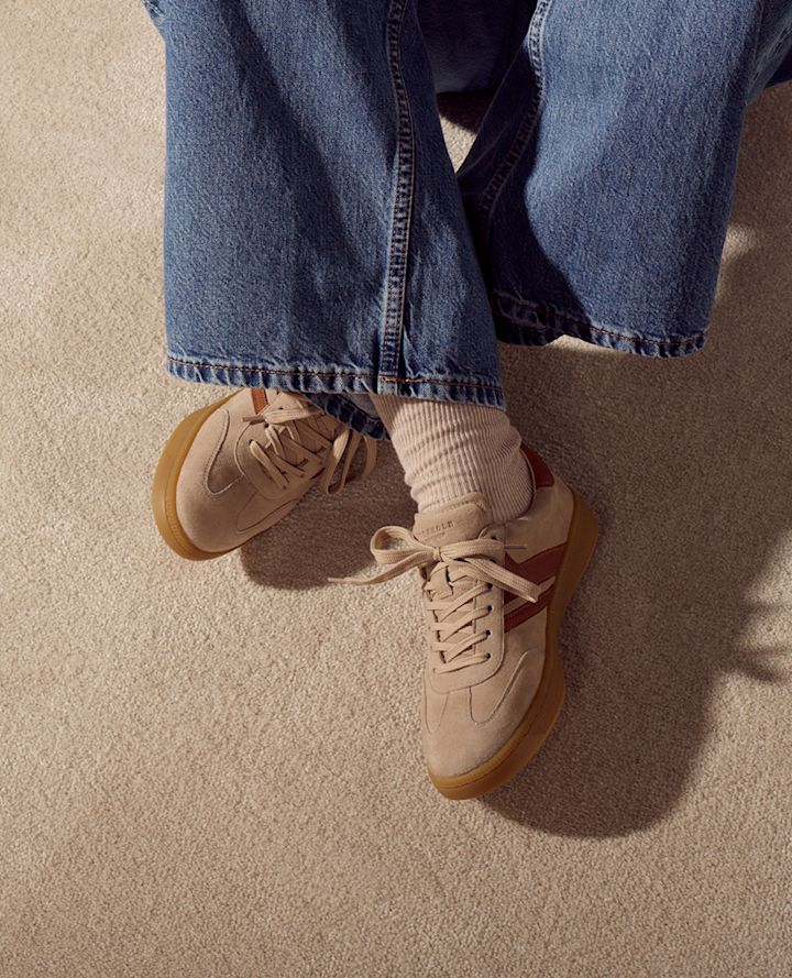 Beige semsket sneakers med gummisåle og røde detaljer, brukt med vide jeans og hvite sokker på beige teppe.