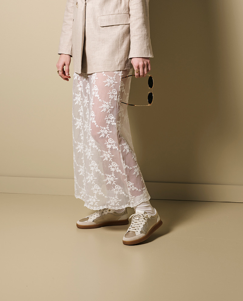 Person i beige kavaj och transparent spetskjol med vita blommönster, bär beige-vita sneakers med gummisula och håller solglasögon.