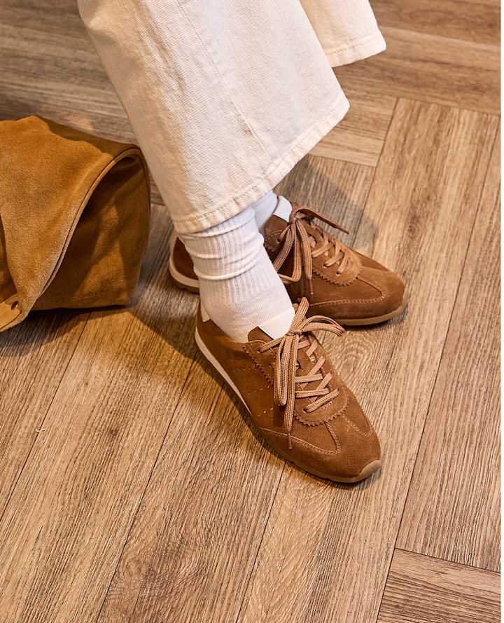 Bruna sneakers i mocka med snörning, retrostil. Bärs med vita strumpor och beige byxor på ett trägolv.