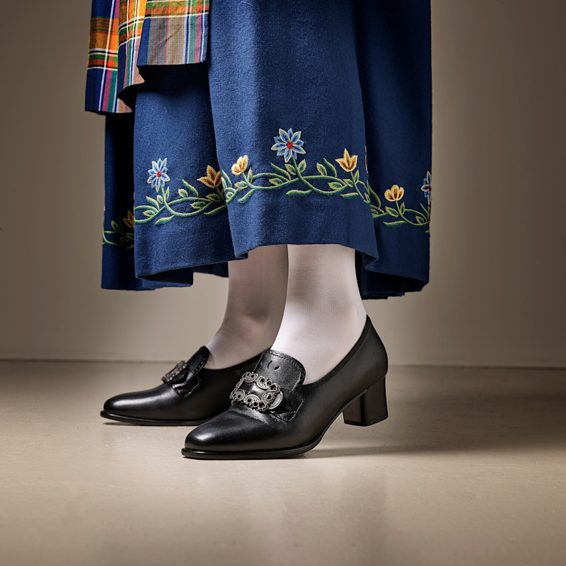 Sorte sko med lav hæl og dekorativ spenne på føttene til en person iført hvite strømper og blå bunadskjørt med broderte blomster nederst.