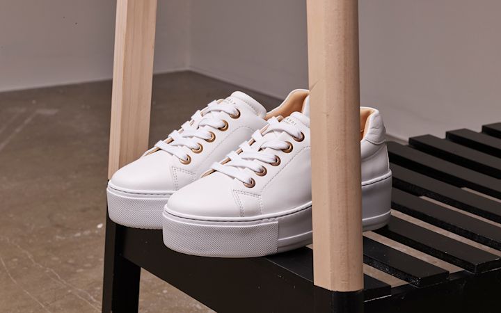 Vita sneakers med tjocka sulor placerade på en svart bänk med ljusa träben i ett rum med betonggolv.