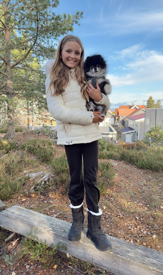 Flicka i vit jacka och svarta byxor håller en liten svartvit hund ute i ett skogsområde med hus i bakgrunden.