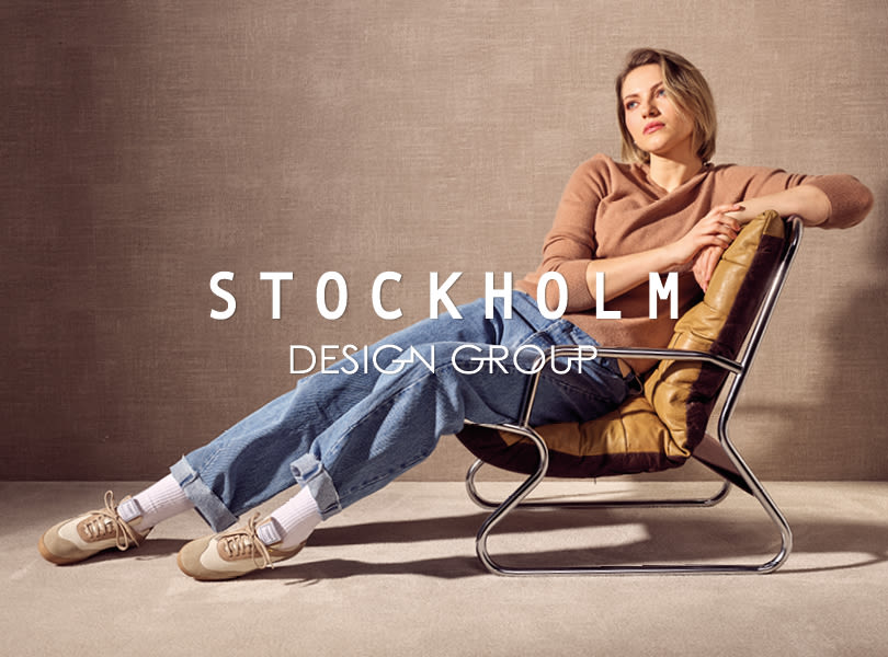 Modell i lenestol med beige sneakers fra Stockholm Design Group, brun genser og jeans. Stockholm Design Group-logo vises i forgrunnen.