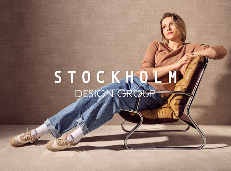 Person i beige tröja och jeans sitter i designfåtölj, bär beiga sneakers med vita strumpor. Stockholm Design Group-logotyp syns.