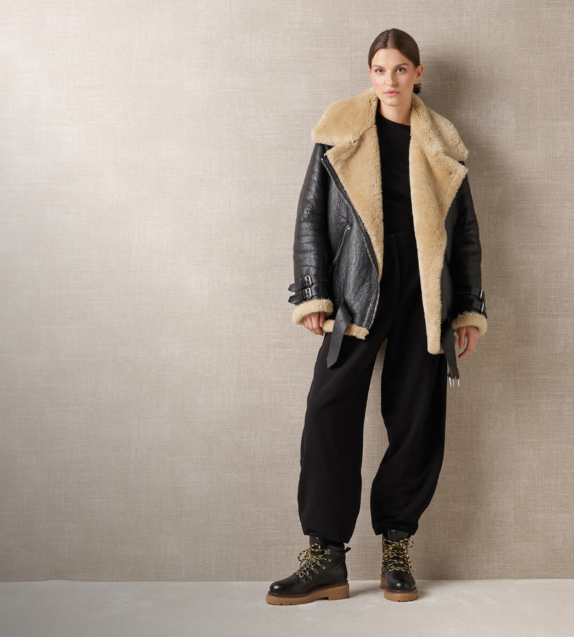 Modell i svart antrekk med skinnjakke med beige pelskrage og svarte boots fra Stockholm Design Group med gule lisser og brun såle.