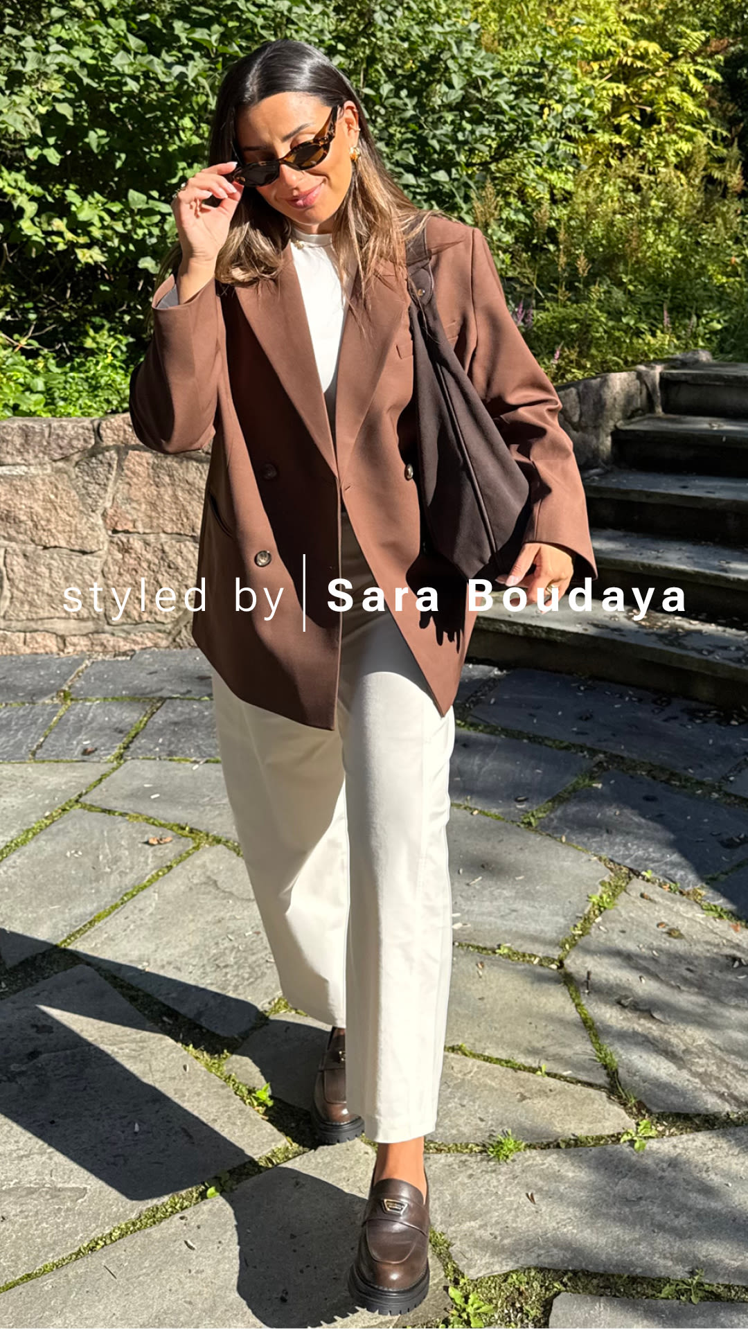 Sara Boudaya i brun blazer og hvite bukser med brune chunky loafers i skinn fra Stockholm Design Group, står utendørs i solskinn med solbriller.