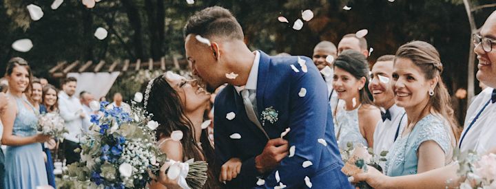 Brud och brudgum kysser omgivna av brudfölje som ler och kastar vita blomblad utomhus i en trädgård.