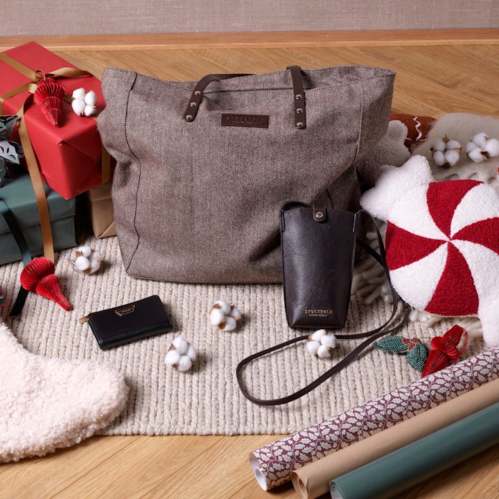Brun tweedväska med läderdetaljer omgiven av julklappar, plånbok och mobilfodral på en stickad matta med bomullsdekorationer.
