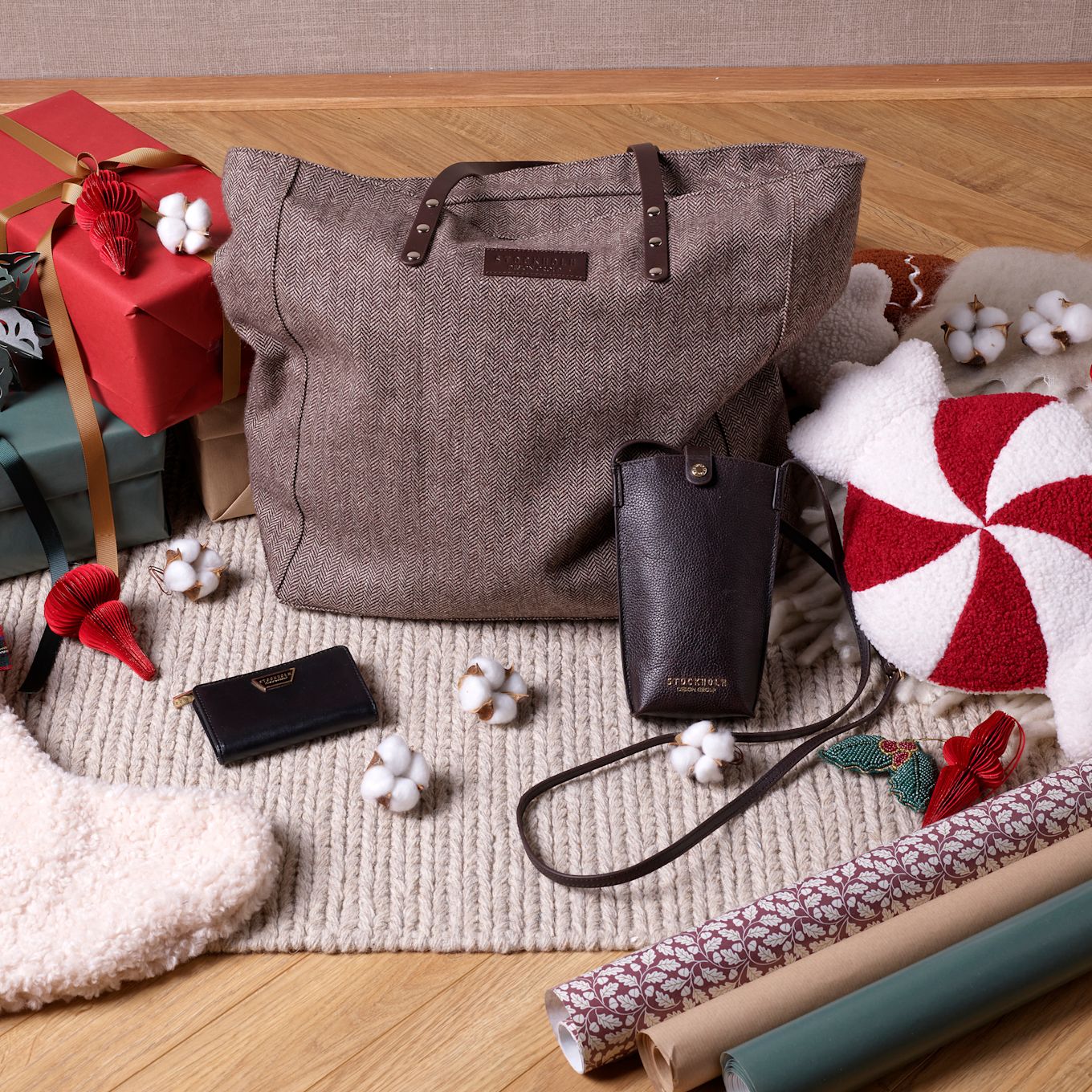 Brun tweedväska med läderdetaljer omgiven av julklappar, plånbok och mobilfodral på en stickad matta med bomullsdekorationer.