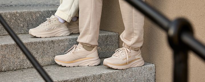 Två personer sitter på en granittrappa med beige joggingskor och ljusbeige byxor, nära en vägg med svart räcke i förgrunden.