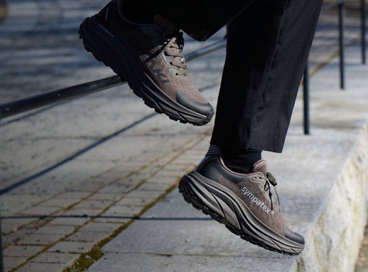 Person i mörka byxor bär bruna Okaki Sympatex vandringsskor med tjock sula, tar ett steg på stenbelagd yta.