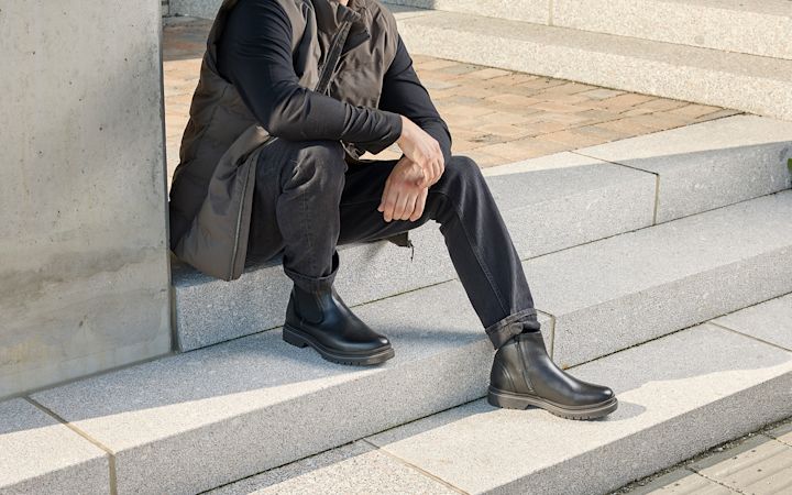 Mann sitter på utendørs betongtrapp iført svart genser, grå vest, svarte jeans og svarte skinnstøvler.