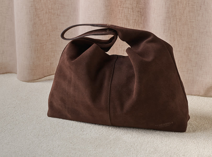 Elegant mørkebrun semsket skinnveske med håndtak fra Stockholm Design Group, plassert på et lyst teppe mot en kremfarget gardinbakgrunn.