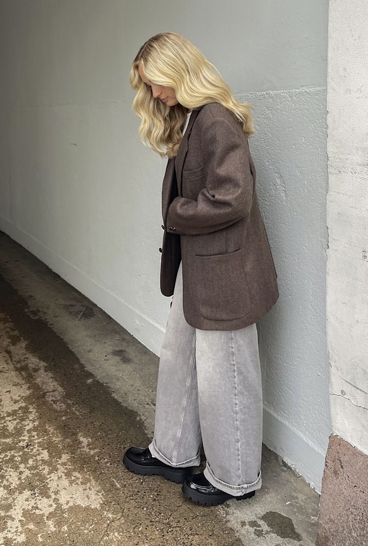 Blont kvinna som bär en brun kavaj, ljusgrå vida byxor och svarta klumpiga skor står vid en grå vägg.