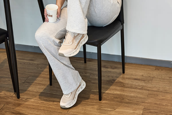 Person i gråa mjukisbyxor sitter på svart stol med kaffemugg, bär beiga sneakers Okaki Mile i sportig stil med tjock sula.