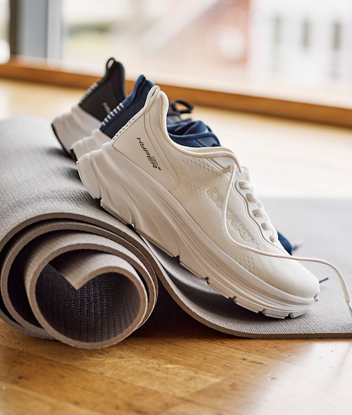 Hvite og mørkeblå Okaki Hyper med tykk såle plassert på en rullet yogamatte, moderne og sporty design.