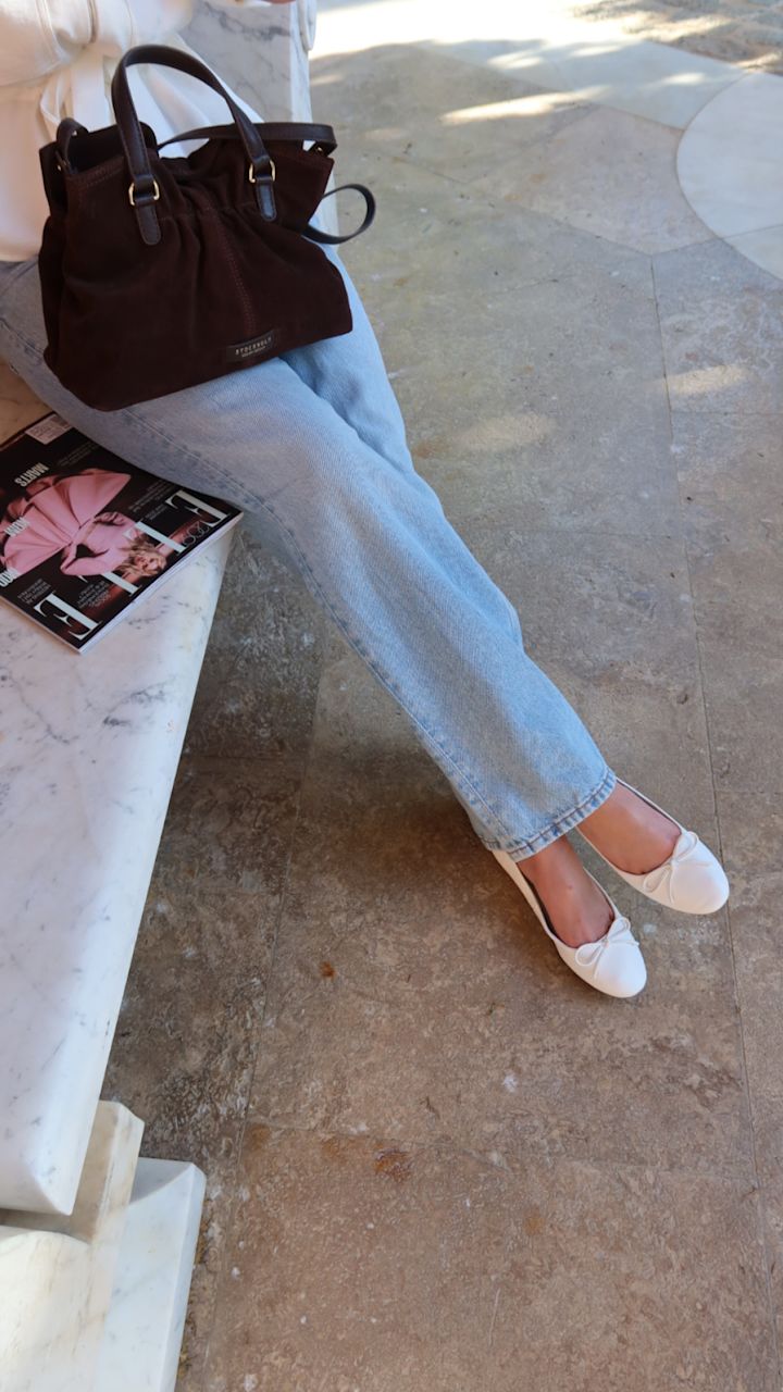 Person i lyseblå jeans med hvite ballerinasko fra Privé og brun semsket veske fra Stockholm Design Group sittende ved et marmorbord med et magasin.