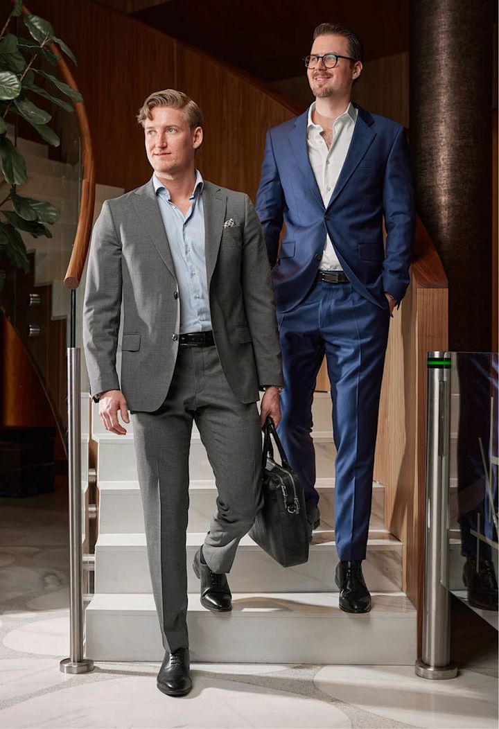 To menn i dress, en i grå og en i blå, går ned en trapp i et moderne kontormiljø.