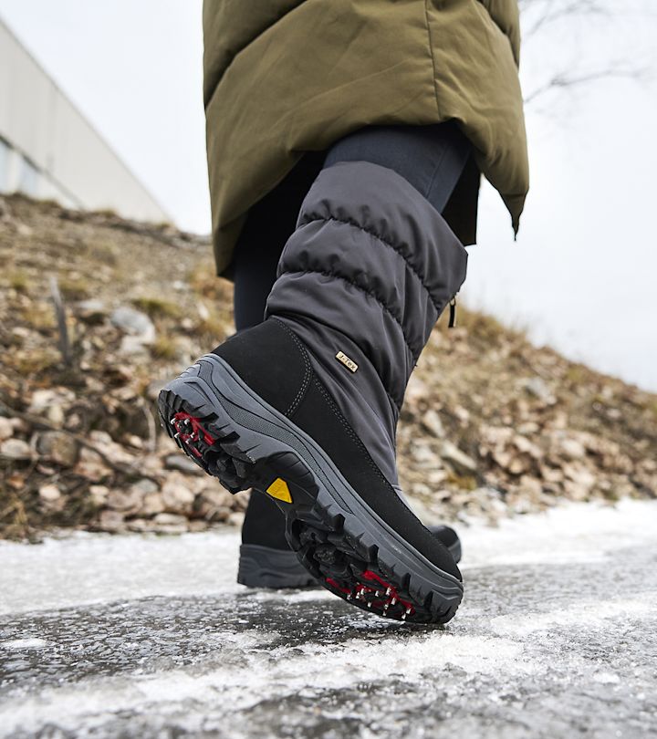 Vinterstövel med dubbar på sulan som går på isig väg, iklädd mörka byxor och grön jacka utomhus.