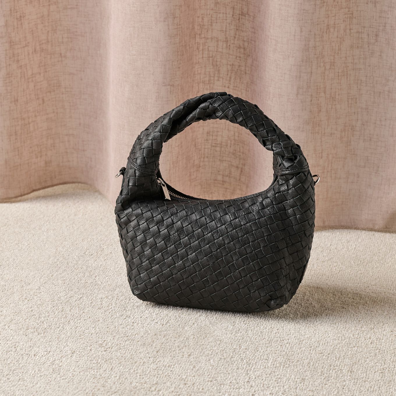 Svart flettet skinnveske med håndtak plassert på beige teppe mot en lys bakgrunn.