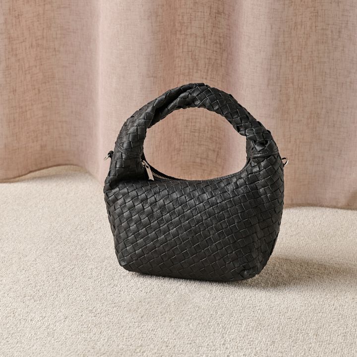 Svart flettet skinnveske med håndtak plassert på beige teppe mot en lys bakgrunn.