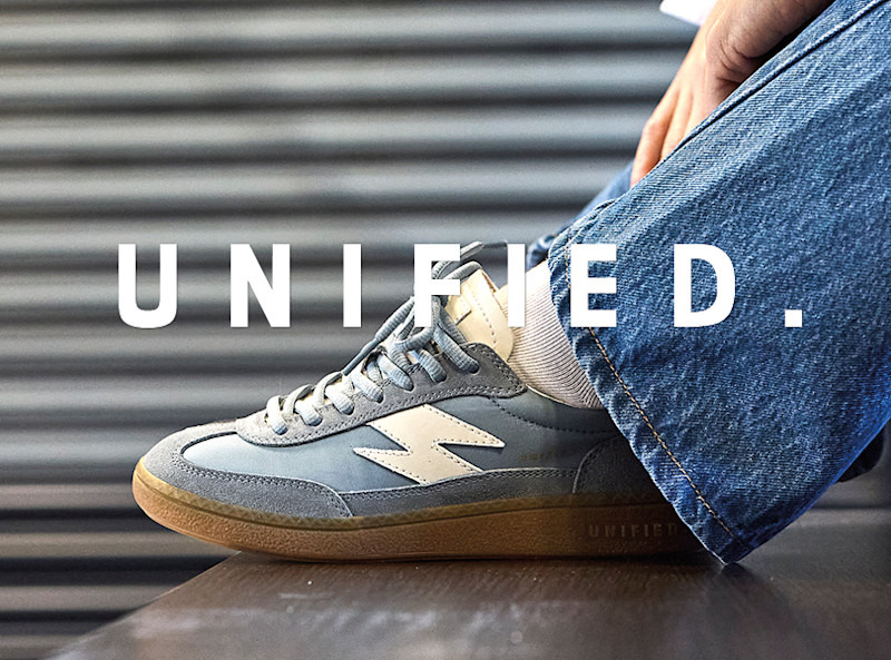 Lyseblå sneakers med hvite detaljer, brukt med jeans. Logoen "UNIFIED." vises over skoen mot en metallisk bakgrunn.