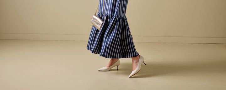 Kvinne iført blå og hvitstripet kjole og hvite høye hæler som går på beige gulv med liten gullfarget veske hengende over skulderen.