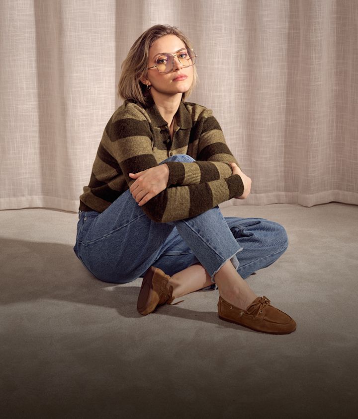 Person i randig tröja och jeans sitter på golv med bruna mockasiner, stilrena seglarskor från Stockholm Design Group med snören i klassisk design.