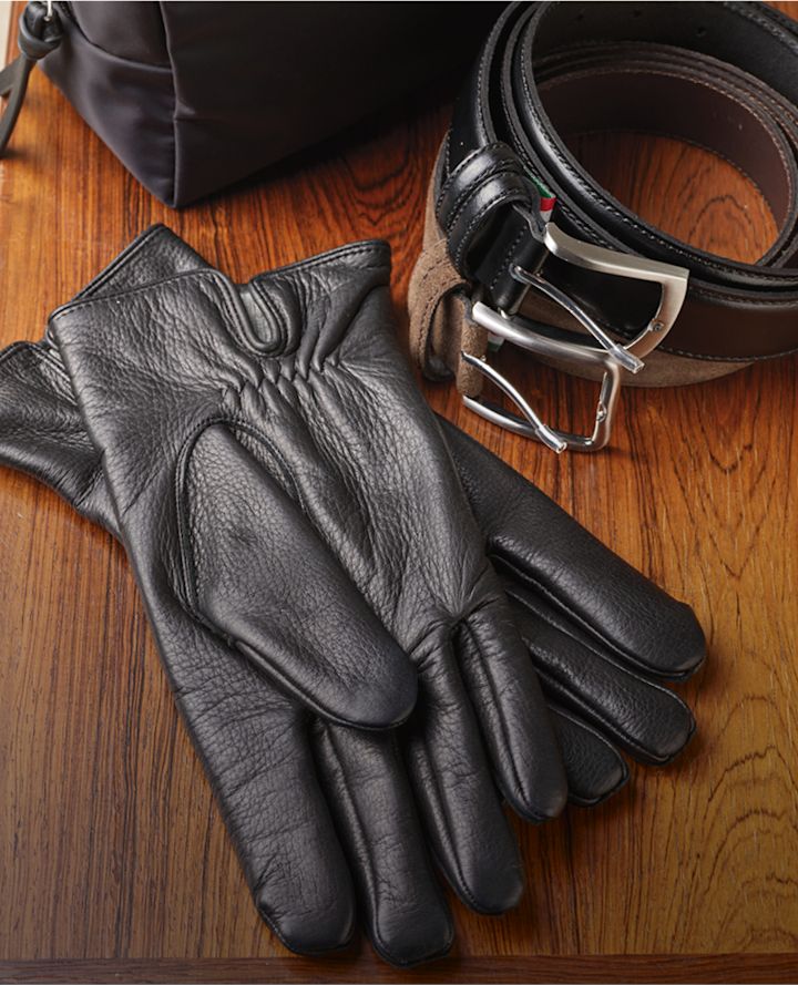 Svarta läderhandskar från Stockholm Design Group, ett svart och brunt läderbälte med metallspänne och en liten svart väska på träyta.