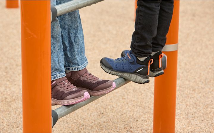 To barn står på en metallstang i en lekeplass med oransje stolper, iført blå og lilla sko.