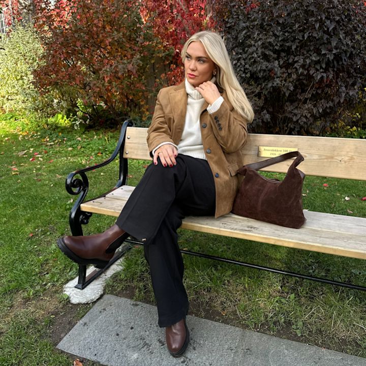 Kvinna med blont hår sitter på en bänk i parken, iklädd en brun jacka, vit tröja, svarta byxor och bruna stövlar, med en brun väska bredvid sig.