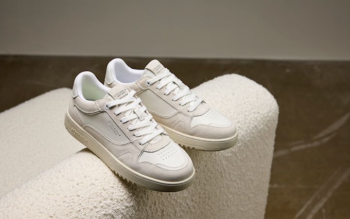 Vita sneakers med skinn- och mockadetaljer placerade på en krämfärgad, texturerad yta i ett neutralt rum.
