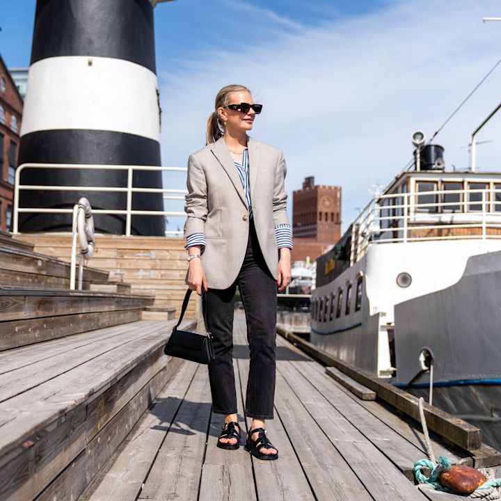 Person i grå blazer på brygge med båter, iført svarte åpne sandaler med remmer i sporty stil.