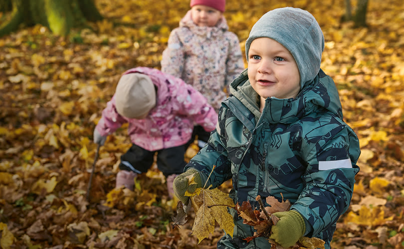 Småbarn i färgglada vinterkläder leker bland höstlöv. Barnet i förgrunden bär grön jacka och mössa.