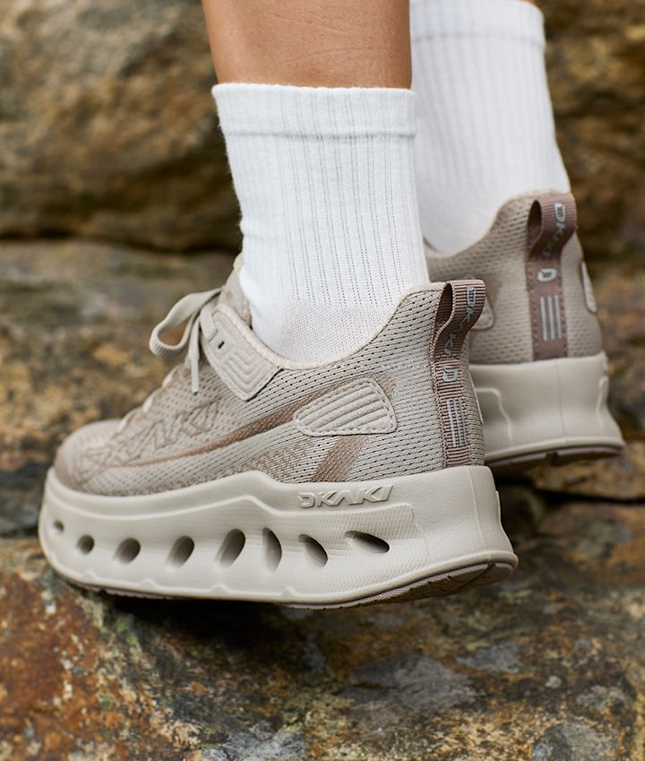 Beige fritidssko fra Okaki med hvite sokker, vist på steingrunn. Skoene har perforert såle og nettingmønster.