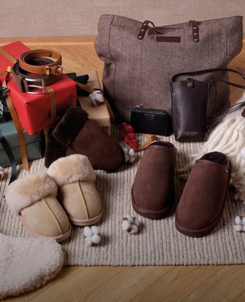 Mysiga tofflor i beige och brun mocka med fårskinnsfoder, bredvid en grå väska, plånbok och presentaskar på en stickad matta.