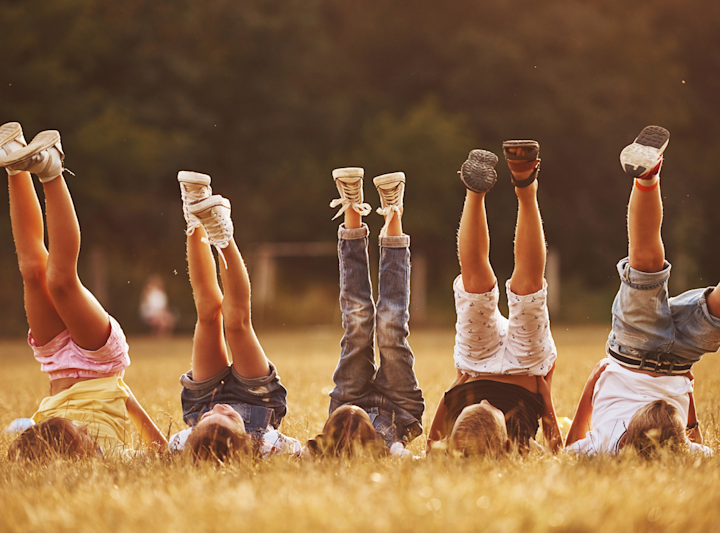 Fem barn ligger på rygg i en äng med benen utsträckta upp mot himlen en solig dag.